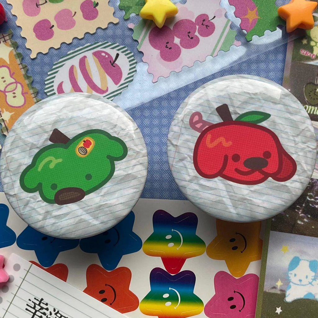Two round pinback buttons with cute cartoon designs, one featuring a green apple with a rainbow and the other a red apple with a black nose, displayed on a colorful collage background.