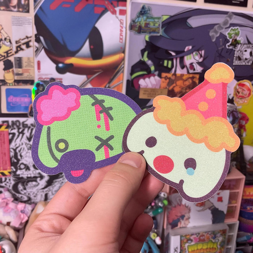 Zombie and clown dog stickers in dog head shape, one green with pink puff on head and one with clown hat, tear and red nose held by a hand
